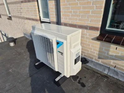 Daikin 4 kW warmtepomp op bigfoot opstelbalken op een  houten dak van een uitbouw, naast een stenen mu